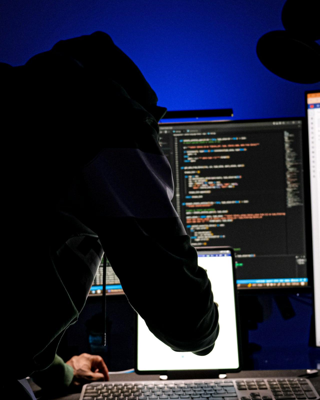 A hooded figure hunched over a keyboard in front of monitors full of code, lit in blue.