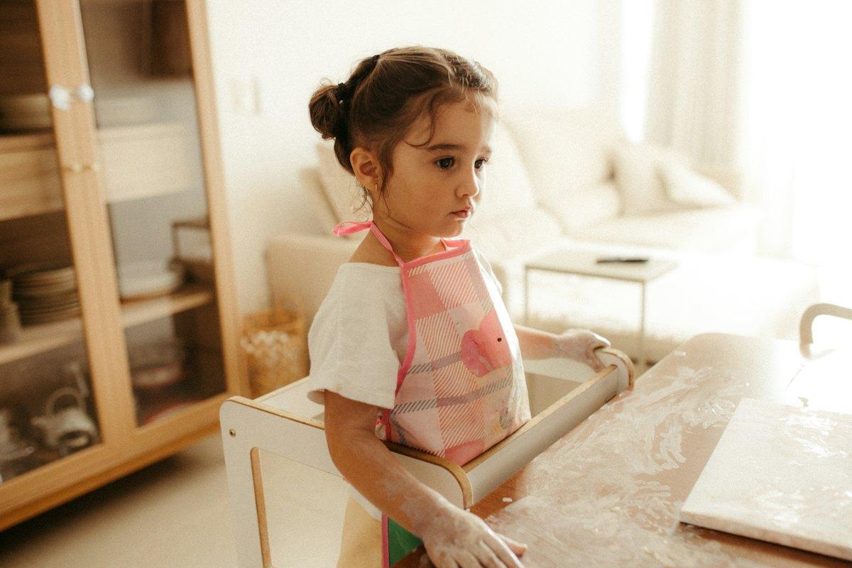 A young girl in an apron stands at a kitchen table dusted with flour.
