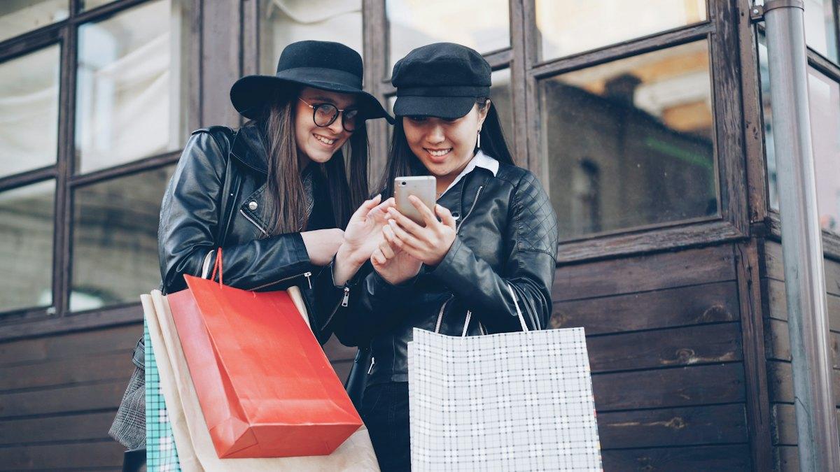 Two friends with shopping bags look at a phone together.