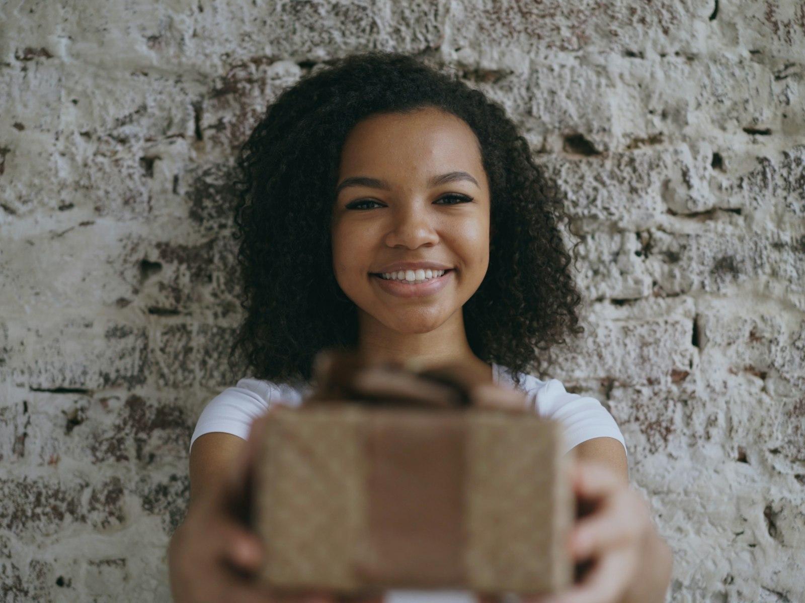 A smiling young Black woman holding out a wrapped gift box